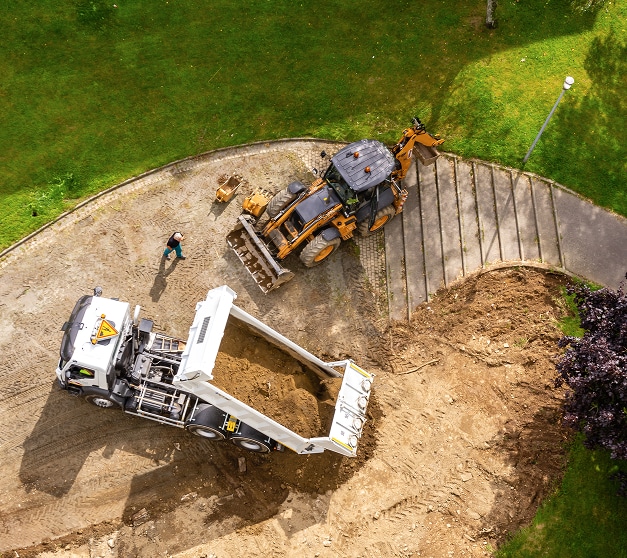 Chantier de terrassement : vue aérienne d'un camion benne blanc et d'une rétrocaveuse Case. Un ouvrier supervise le chargement de la terre.